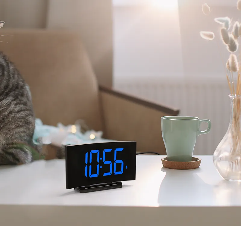 Curved alarm clock with blue digits on a white table by a window